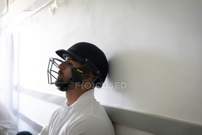 Vista lateral de perto de um jogador de críquete misto de raça masculina vestindo brancos e capacete de críquete, sentado em um banco em um vestiário, inclinando a cabeça na parede. — Fotografia de Stock