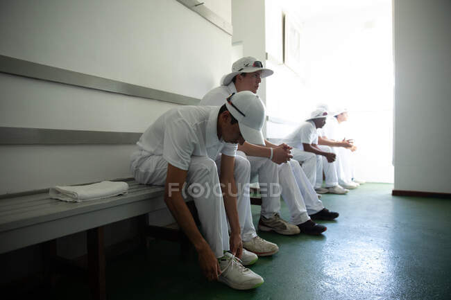 Vista lateral de un equipo de jugadores de cricket masculinos multiétnicos adolescentes que usan blancos, sentados en un banco en un vestuario, preparándose para el juego, uno de los jugadores está atando sus cordones de zapatos. - foto de stock