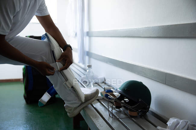 Vue de côté section basse du joueur de cricket masculin portant des blancs, debout dans un vestiaire, se préparant au jeu, mettant sur un garde-jambes. — Photo de stock