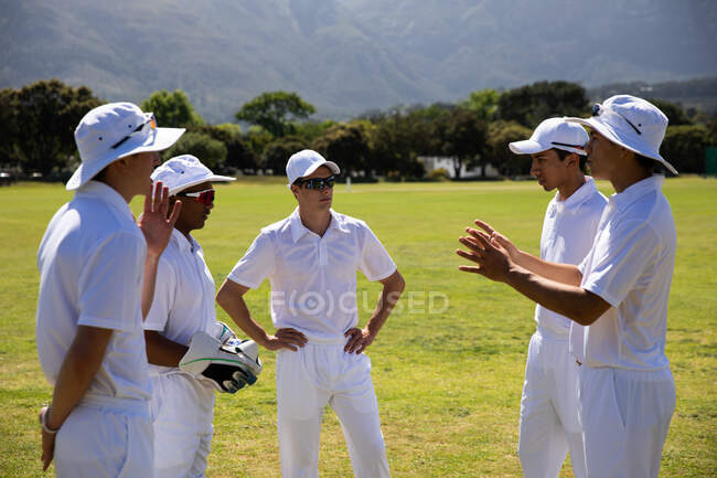 Vista lateral de un grupo de jugadores de cricket masculinos multiétnicos adolescentes que usan blancos, parados en un campo de cricket, discutiendo el juego durante un día soleado. - foto de stock