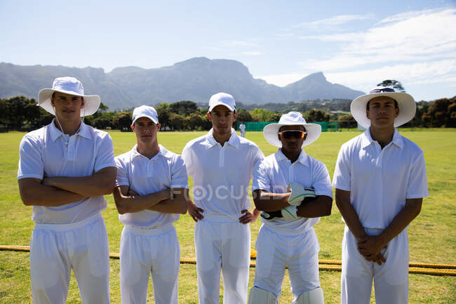 Передний вид подростковой многонациональной мужской команды по крикету в белых одеждах, стоящей на поле вместе со скрещенными руками, смотрящей прямо в камеру — стоковое фото
