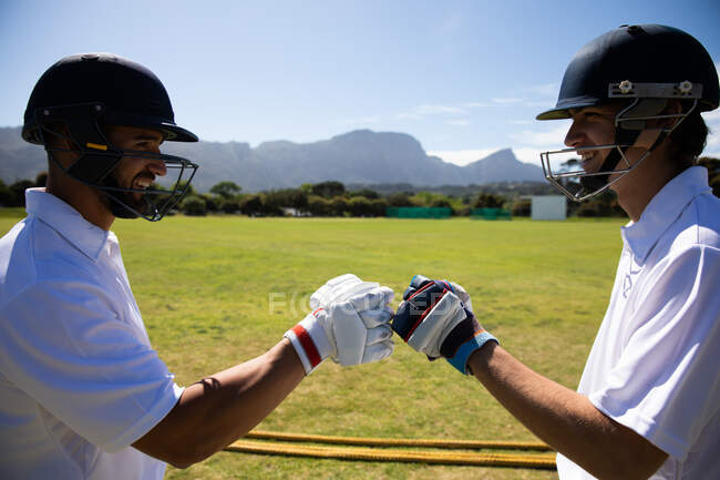 Vista lateral de dois jogadores de críquete masculinos multi-étnicos adolescentes vestindo brancos e capacetes de críquete, em pé no campo, punho-batendo e sorrindo um para o outro. — Fotografia de Stock