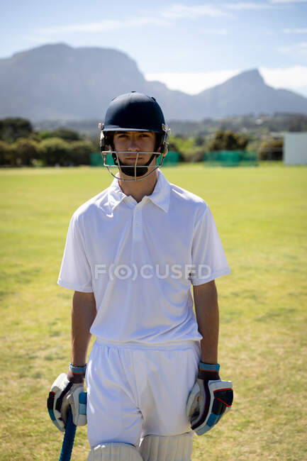Ritratto di un adolescente caucasico fiducioso giocatore di cricket che indossa bianchi da cricket, casco e guanti da cricket, in piedi su un campo da cricket in una giornata di sole guardando alla fotocamera — Foto stock