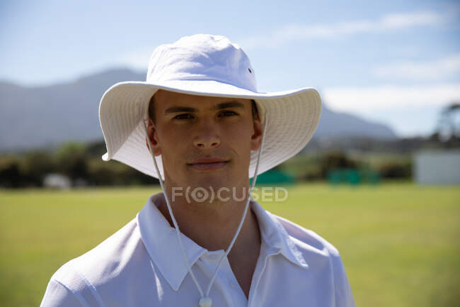 Porträt eines selbstbewussten jungen kaukasischen Cricketspielers mit weißem Cricket und breitem Hut, der an einem sonnigen Tag auf einem Cricketplatz steht und in die Kamera blickt — Stockfoto