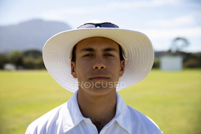 Portrait of a confident teenage Caucasian male cricket player wearing cricket whites, and a wide brimmed hat and sunglasses, standing on a cricket pitch on a sunny day looking to camera — Stock Photo