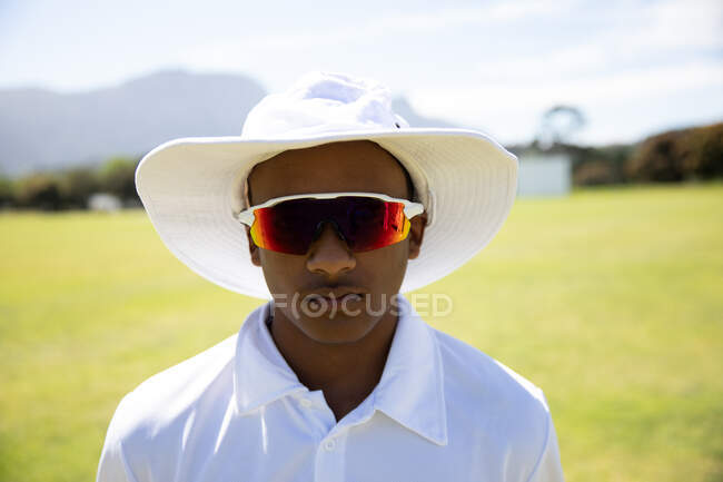Retrato de um adolescente confiante jogador de críquete asiático vestindo branco de críquete, um chapéu de aba larga e óculos de sol, de pé em um campo de críquete em um dia ensolarado olhando para a câmera — Fotografia de Stock