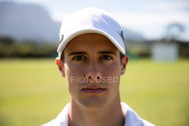 Портрет уверенного подростка, белого игрока в крикет в белом и кепке, стоящего на поле для крикета в солнечный день, смотрящего в камеру. — стоковое фото