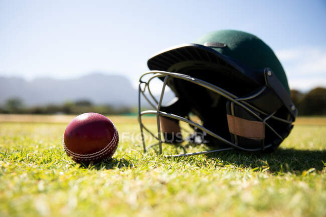 Close up view of a red cricket ball and a green cricket helmet lying on a cricket pitch on a sunny day — Stock Photo
