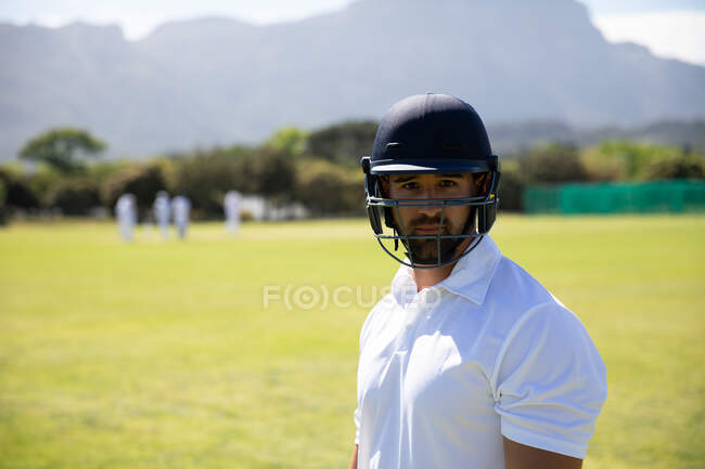 Retrato de un jugador de críquet masculino de raza mixta confiado que usa blancos de críquet y casco, parado en un campo de críquet en un día soleado mirando a la cámara - foto de stock