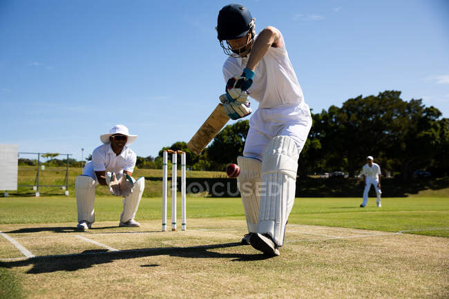 Передній вид підлітка з кавказького крікету, одягненого в шолом і утримуючи крикетний кажан, намагається вдарити по м'ячу під час матчу в крикет, а інші гравці залишаються на задньому плані. — стокове фото
