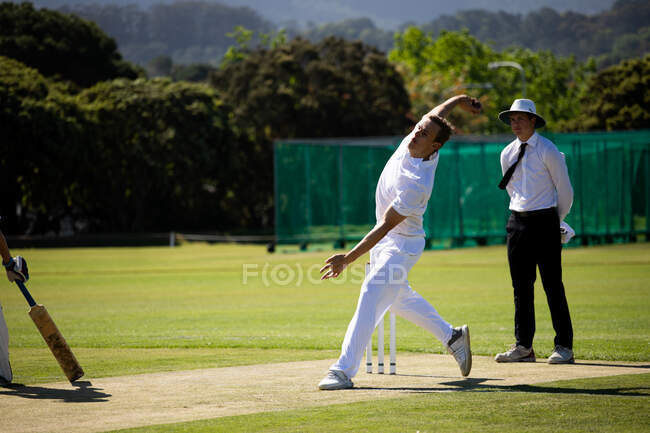 Vista frontale di un adolescente bianco giocatore di cricket che indossa i bianchi, oscillando cercando di lanciare la palla sul campo durante una partita di cricket, con un arbitro in piedi sullo sfondo. — Foto stock