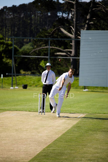 Vista frontal de um adolescente caucasiano jogador de críquete vestindo brancos, jogando a bola em campo durante uma partida de críquete, com um árbitro em pé no fundo. — Fotografia de Stock