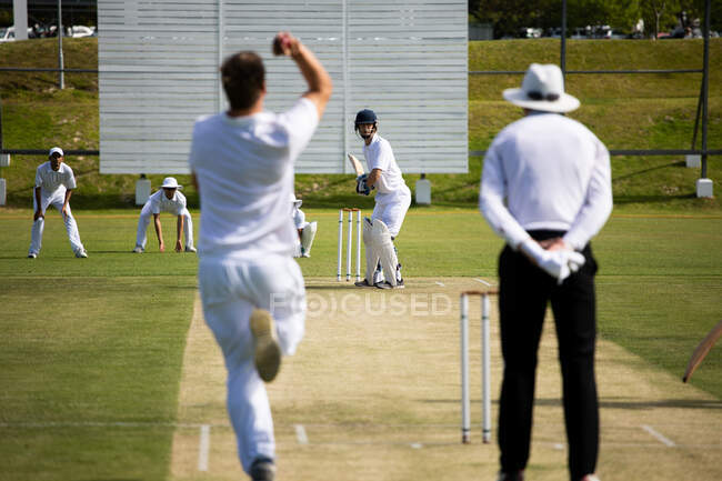 Rear view of a teenage Caucasian male cricket player wearing whites, throwing the ball on the pitch during a cricket match, with an umpire standing behind him. — Stock Photo