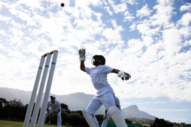 Vue latérale à angle bas d'un adolescent joueur de cricket mixte sur le terrain portant un casque et des gants, essayant d'attraper la balle lors d'un match de cricket, avec d'autres joueurs jouant en arrière-plan. — Photo de stock