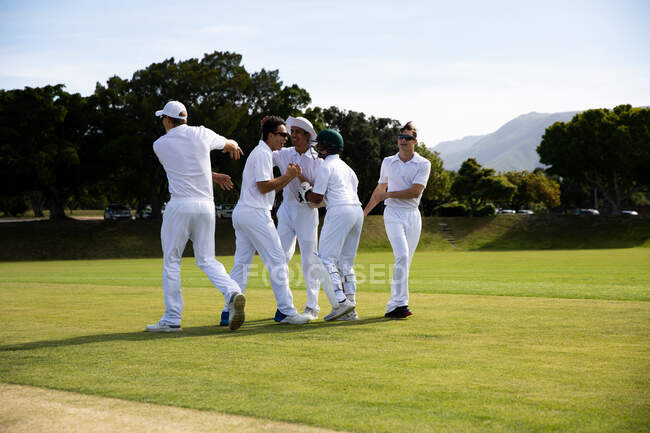 Vista lateral de uma equipe de críquete masculina multiétnica adolescente vestindo brancos, em pé no campo, abraçando, sorrindo e conversando durante o dia ensolarado. — Fotografia de Stock