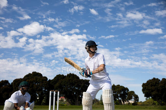 Vista frontal de un jugador de cricket masculino caucásico adolescente en el campo con casco y guantes, sosteniendo un bate de cricket, tratando de golpear la pelota durante un partido de cricket, con otro jugador en cuclillas en el fondo.. - foto de stock