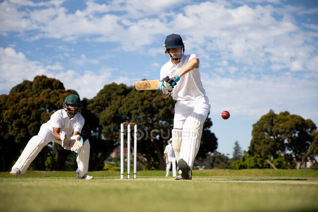 Vista frontal de un jugador de cricket masculino caucásico adolescente de pie en el campo con casco y guantes, sosteniendo un bate de cricket, golpeando la pelota durante un partido de cricket, con otro jugador listo para atrapar una pelota en el fondo. - foto de stock