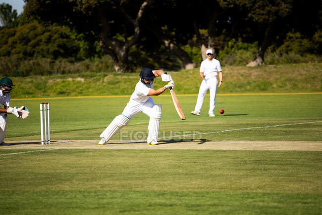 Vista frontal de um adolescente caucasiano jogador de críquete em pé no campo usando capacete e luvas, segurando um taco de críquete, batendo a bola durante uma partida de críquete, com outro jogador em pé no fundo. — Fotografia de Stock