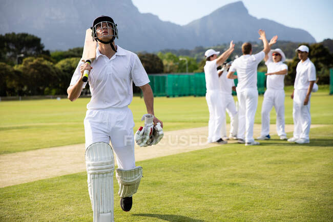 Вид спереди на подростка, белого игрока в крикет в белых шлемах, идущего через поле, держащего биту для крикета и перчатки, в то время как другие игроки сжимаются на заднем плане. — стоковое фото