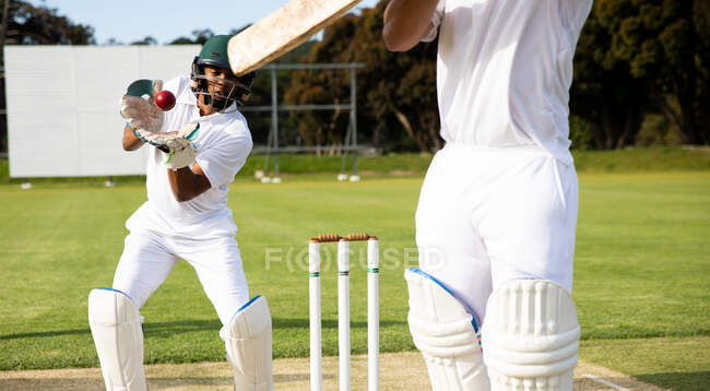 Вид спереди юношеского белого игрока в крикет на поле во время матча по крикету, ловящего мяч с другим игроком, держащим в передней части крикетную биту — стоковое фото