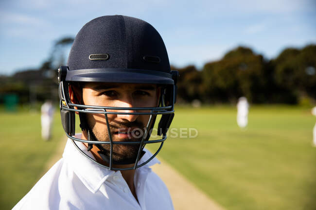 Retrato de um jogador de críquete misto confiante vestindo branco de críquete e capacete, de pé em um campo de críquete em um dia ensolarado olhando para a câmera com outros jogadores em pé no fundo. — Fotografia de Stock