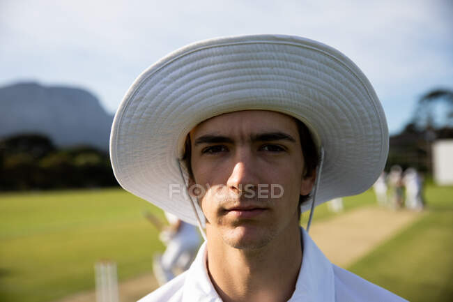 Портрет уверенного молодого белого игрока в крикет в белых крикетных шапках и широкой шляпе, стоящего на поле для крикета в солнечный день, смотрящего в камеру, с другими игроками, стоящими на заднем плане. — стоковое фото