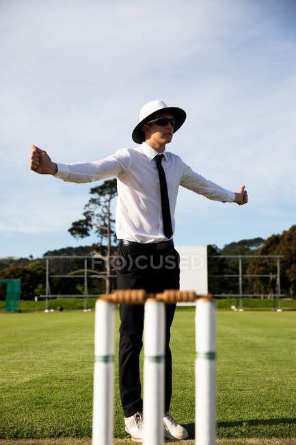 Vista lateral de um árbitro de críquete masculino caucasiano vestindo camisa branca, gravata preta, um chapéu de aba larga e óculos de sol, de pé em um campo de críquete pelo postigo, espalhando as mãos com os polegares para cima. — Fotografia de Stock