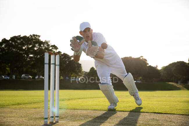 Вид спереди на подростка-белого игрока в крикет, одетого в белое и стаканчик, пытающегося поймать мяч для крикета, калиткой на поле в солнечный день — стоковое фото