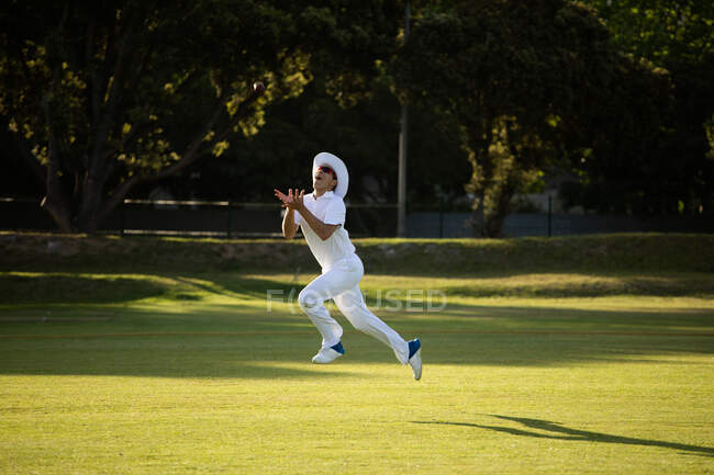 Vista lateral de um adolescente de raça mista jogador de críquete vestindo brancos e um chapéu, pulando para cima tentando pegar uma bola de críquete em campo durante um dia ensolarado — Fotografia de Stock