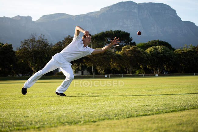 Побочный обзор подростка-белого игрока в крикет в белых одеждах, прыгающего, пытаясь поймать мяч для крикета на поле в солнечный день — стоковое фото