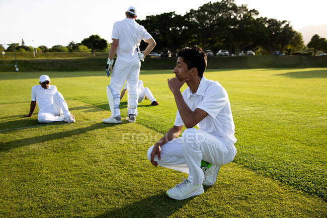 Вид сбоку на юношескую многонациональную команду по крикету, носящую белые, сидящую или стоящую на коленях на поле, отдыхающую после матча. — стоковое фото