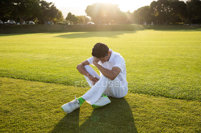 Vista frontale di un adolescente triste giocatore di cricket maschile di razza mista che indossa i bianchi, seduto sul campo, a riposo dopo la partita durante una giornata di sole. — Foto stock