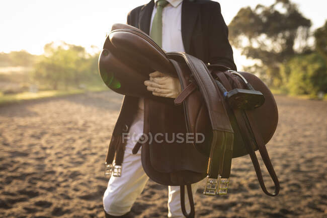 Vista frontal sección central de un jinete de caballos macho vestido con elegancia, de pie en un paddock sosteniendo una silla de montar marrón en un día soleado. - foto de stock