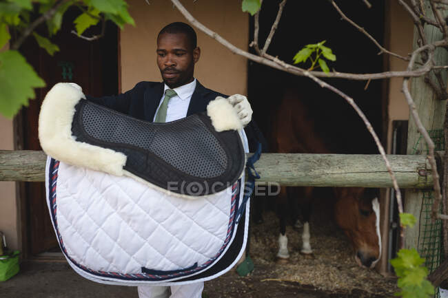 Front view of a smartly dressed African American man preparing to saddle the horse before dressage horse riding, standing by a horse stable — Stock Photo