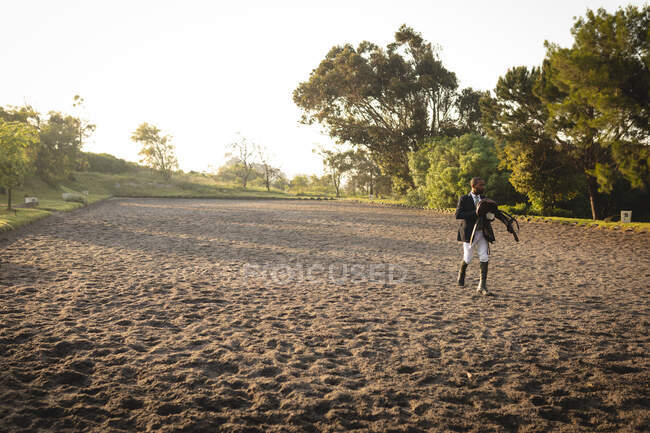 Vista frontale di un cavaliere afroamericano vestito con eleganza che attraversa un paddock portando una sella in una giornata di sole. — Foto stock