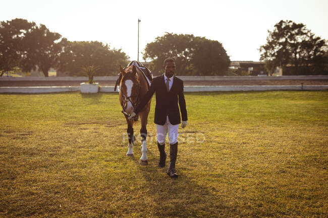 Vista frontale di un uomo afroamericano elegantemente vestito che cammina su un cavallo di castagno su un paddock prima di andare a cavallo di dressage durante una giornata di sole. — Foto stock