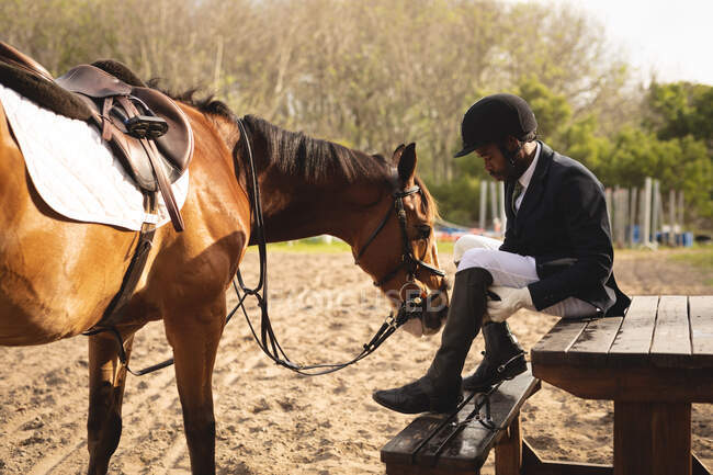 Вид сбоку на ловко одетого афроамериканца, надевающего сапоги перед выездной лошадью в солнечный день, с каштановой лошадью, стоящей рядом с ним на загоне. — стоковое фото