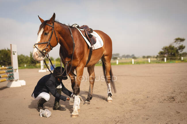 Vista lateral de um homem afro-americano inteligentemente vestido verificando o guarda de tornozelo em seu cavalo castanho antes de um evento de salto de show durante um dia ensolarado . — Fotografia de Stock