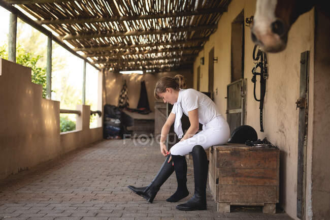 Vue latérale d'une cavalière caucasienne bien habillée se préparant à une compétition de dressage par une journée ensoleillée, enfilant ses bottes d'équitation, tandis que son cheval châtain se tient dans une écurie — Photo de stock