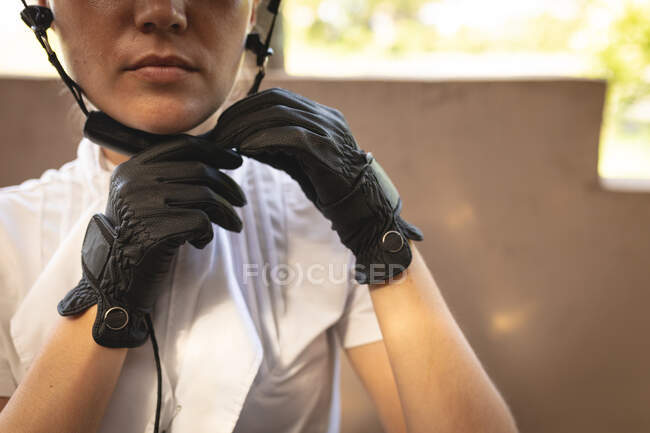 Side view close up of a casually dressed Caucasian female rider wearing black leather gloves, putting on a riding hat during a sunny day. — Stock Photo