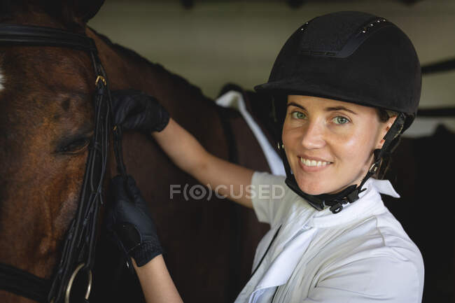 Vista lateral de perto de uma mulher caucasiana casualmente vestida usando um chapéu de equitação, colocando um freio em uma cabeça de cavalo castanha, e olhando para a câmera. — Fotografia de Stock