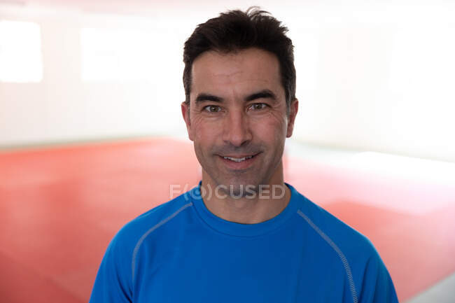 Портрет щасливої змішаної раси чоловічий тренер дзюдо в синій футболці, стоячи в спортзалі, посміхаючись і дивлячись прямо в камеру . — стокове фото