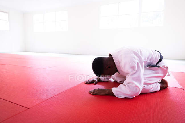Vista lateral de uma judoca masculina de raça mista adolescente vestindo judogi branco, ajoelhado e curvando-se em esteiras no ginásio antes do treinamento de judô. — Fotografia de Stock