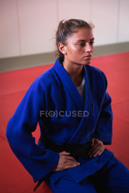 Vue de face gros plan d'une adolescente caucasienne joueuse de judo portant du judogi bleu, agenouillée sur des nattes dans la salle de gym avant l'entraînement de judo. — Photo de stock