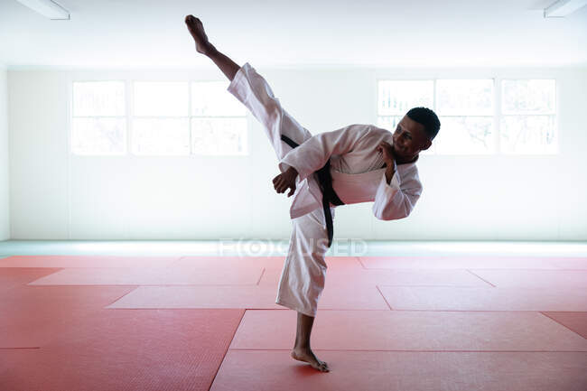 Vista frontale di un adolescente maschio di razza mista che indossa judogi bianchi, si scalda prima di un allenamento in palestra, colpisce una posa, allunga la gamba e calcia l'aria. — Foto stock