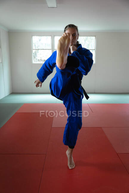 Vista frontal close-up de uma judoca feminina de raça mista adolescente vestindo judogi azul, aquecendo antes de um treinamento em um ginásio, marcando uma pose, esticando a perna e chutando o ar. — Fotografia de Stock
