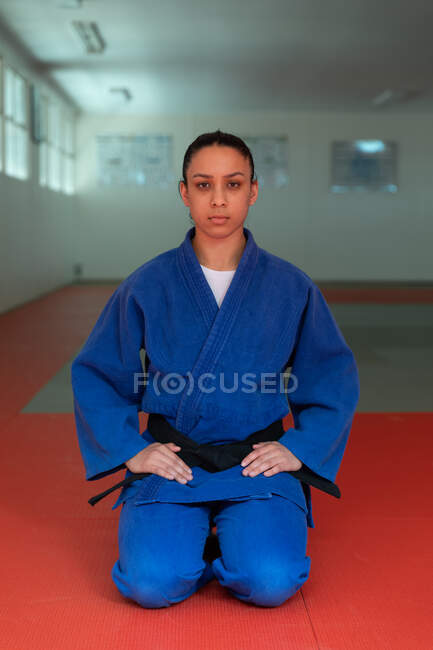 Frontansicht einer selbstbewussten Mixed Race Judoka mit blauem Judogi, die in der Turnhalle auf Matten kniet und direkt in eine Kamera blickt, vor dem Judo-Training. — Stockfoto