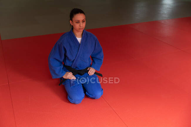 High angle front view of a focused teenage mixed race female judoka wearing blue judogi, kneeling on mats in the gym before judo training. — Stock Photo