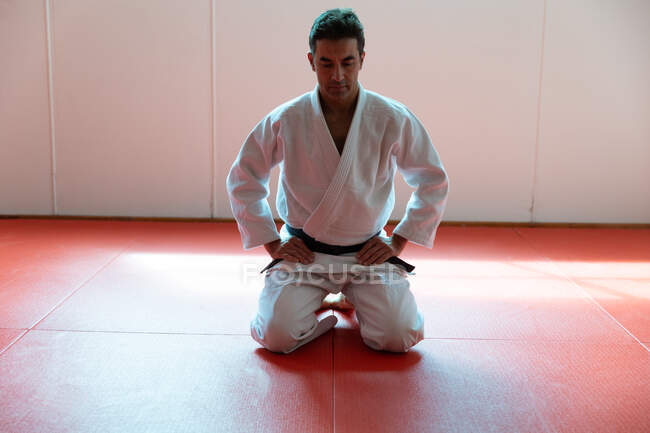 Vue de face d'un entraîneur de judo masculin mixte concentré portant du judogi blanc, agenouillé sur des nattes dans la salle de gym avant l'entraînement de judo. — Photo de stock