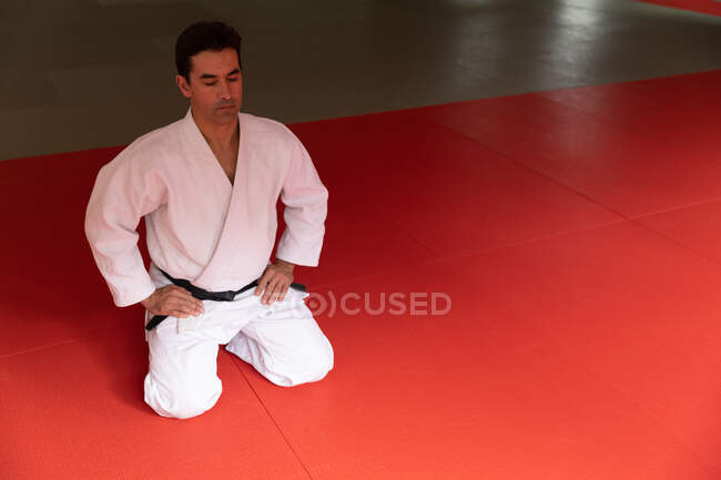 Передній вигляд зосередженої змішаної раси чоловічий тренер дзюдо в білому джудозі, на колінах на килимках у тренажерному залі перед тренуванням дзюдо . — стокове фото